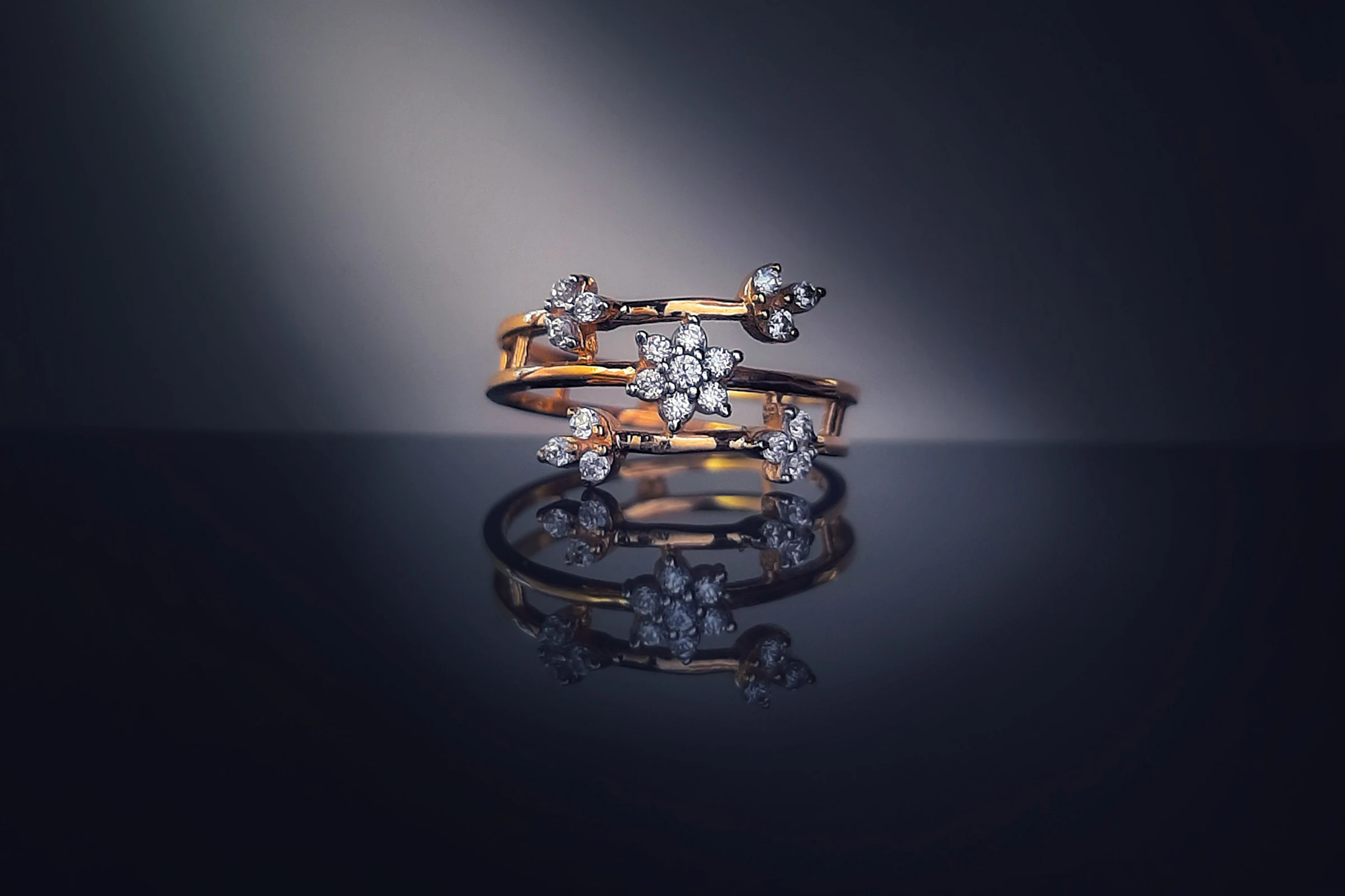 A gold ring set with diamond flowers, spot-lit on a dark mirror